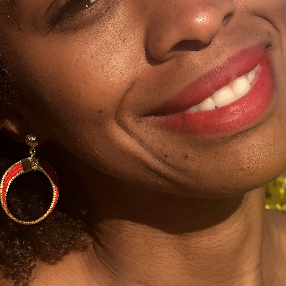 Boucle d'oreille Lola Arco-Íris ORA rouge portée, gros plan sur le sourire, lumière dorée