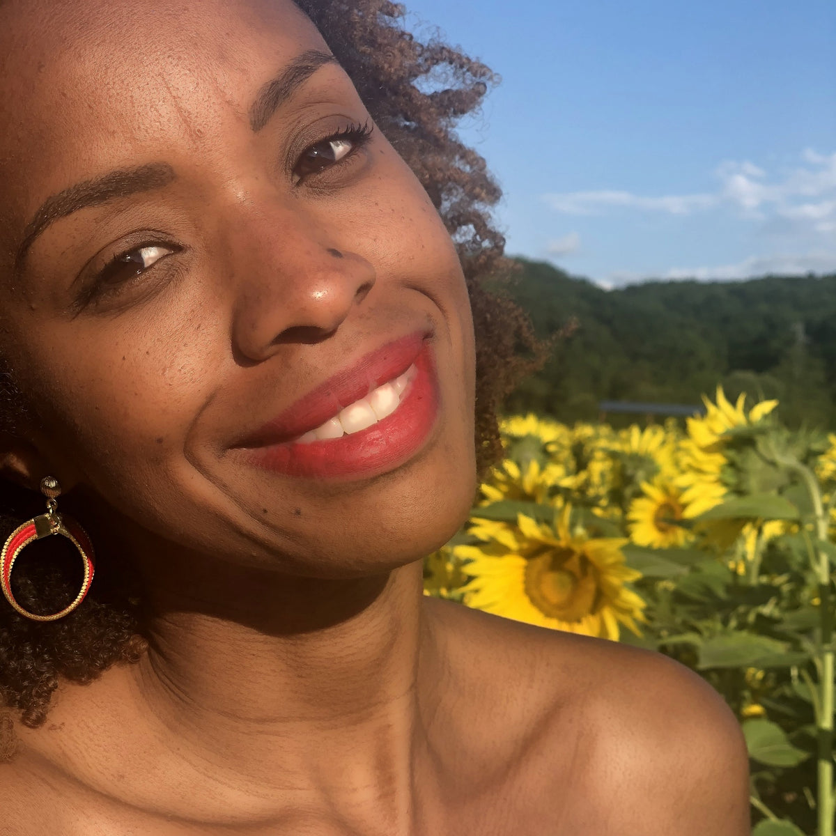 Boucles d'oreilles Lola Arco-Íris ORA rouges portées par Tatiana, champ de tournesols et ciel bleu